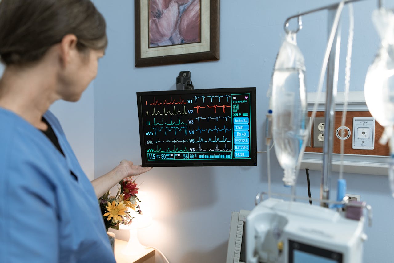 services-03 A nurse in blue scrubs examines a medical monitor displaying vital signs in a hospital setting.